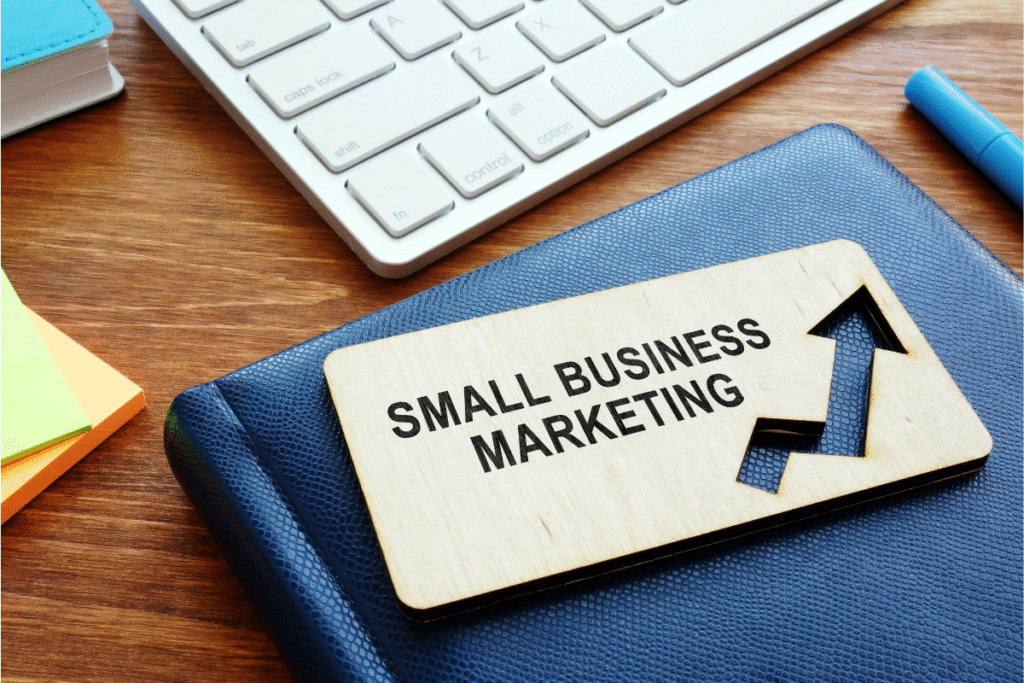 Drewniana tabliczka z napisem „Small Business Marketing” i strzałką symbolizującą wzrost, leżąca na niebieskim notesie przy klawiaturze i kolorowych karteczkach. Grafika symbolizuje marketing dla małych firm.
