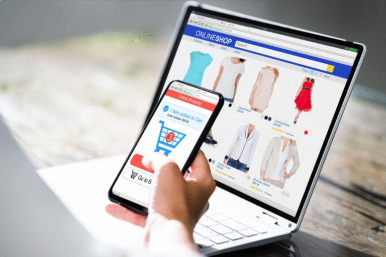 Osoba robiąca zakupy online na laptopie i smartfonie – ekran pokazuje sklep internetowy z odzieżą i dodawanie produktu do koszyka.