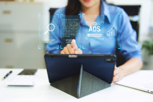 Kobieta obsługująca tablet, nad którym unoszą się cyfrowe ikony reprezentujące feed produktowy: kod XML z danymi produktu, lupa wyszukiwania, napis ADS oraz wykres wzrostu sprzedaży.