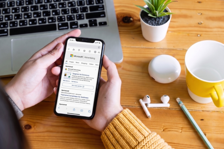 Osoba trzymająca smartfon z wyświetloną reklamą Microsoft Ads na tle laptopa i biurka – reklama Microsoft Ads B2B.